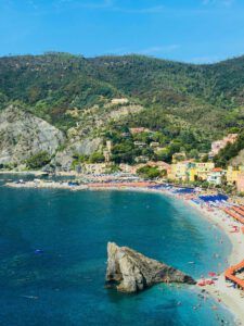 Playa mediterránea con agua turquesa, gran roca en primer plano, sombrillas naranjas y azules y pueblo en la ladera, ideal para ilustrar Los misterios de los 7 años en la Biblia: ¿Qué revela realmente?.