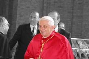 Retrato en color selectivo de un clérigo mayor con vestidura roja y solideo blanco, rodeado de asistentes; ¿Qué dice la Biblia sobre Benedicto XVI?