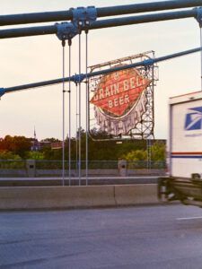 Cartel vintage de Grain Belt Beer visible desde un puente con cables y un camión en movimiento al atardecer — Los 400 años de silencio en la Biblia: ¿Qué sucedió durante ese tiempo?