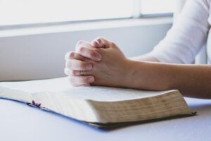 Manos juntas en oración sobre una Biblia abierta junto a una ventana; ¿Qué enseña la Biblia sobre la fe?