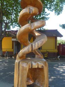 Escultura de madera en espiral tallada al aire libre frente a un edificio amarillo y árboles; ¿Cuándo fue escrito el Génesis de la Biblia?
