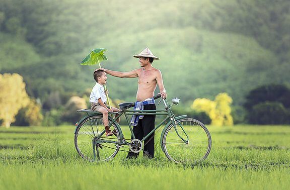Padre sonriente con su hijo en una bicicleta en un campo verde, el hombre usa un sombrero tradicional y sostiene una hoja como paraguas — ¿Qué significa el hijo del hombre en la Biblia? Descubre su verdadero significado aquí