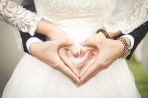 Hasta que la muerte nos separe en la Biblia: ¿Qué dice realmente? Pareja de novios con vestido de encaje y traje formal formando un corazón con las manos sobre el torso.