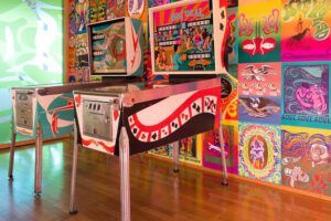 Dos máquinas de pinball retro frente a un muro cubierto de carteles psicodélicos y arte pop: ¿Qué enseña la Biblia sobre el movimiento del 68?